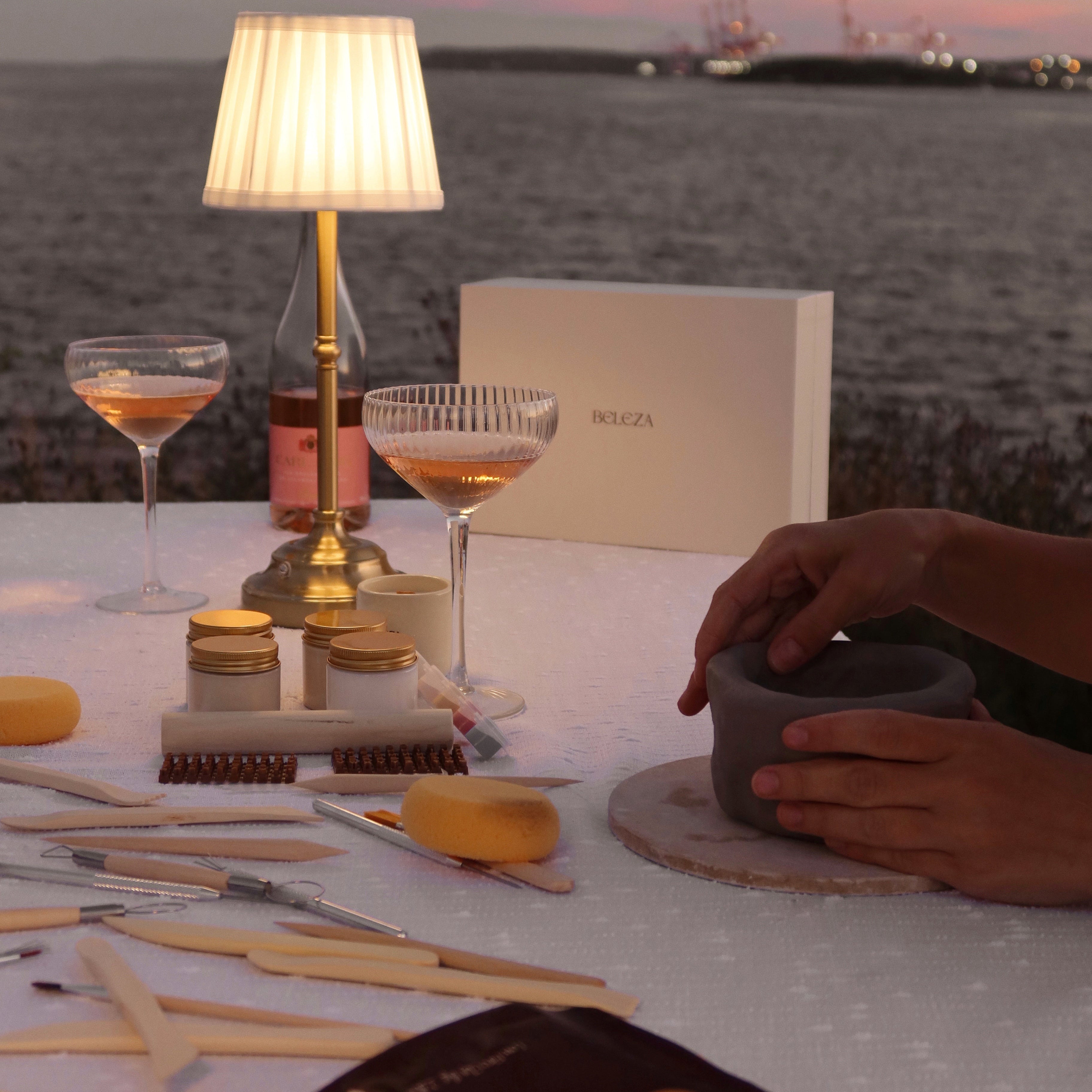 Table setting with drinks and a lamp by a waterfront at dusk whilst making pottery with tools and equipment.